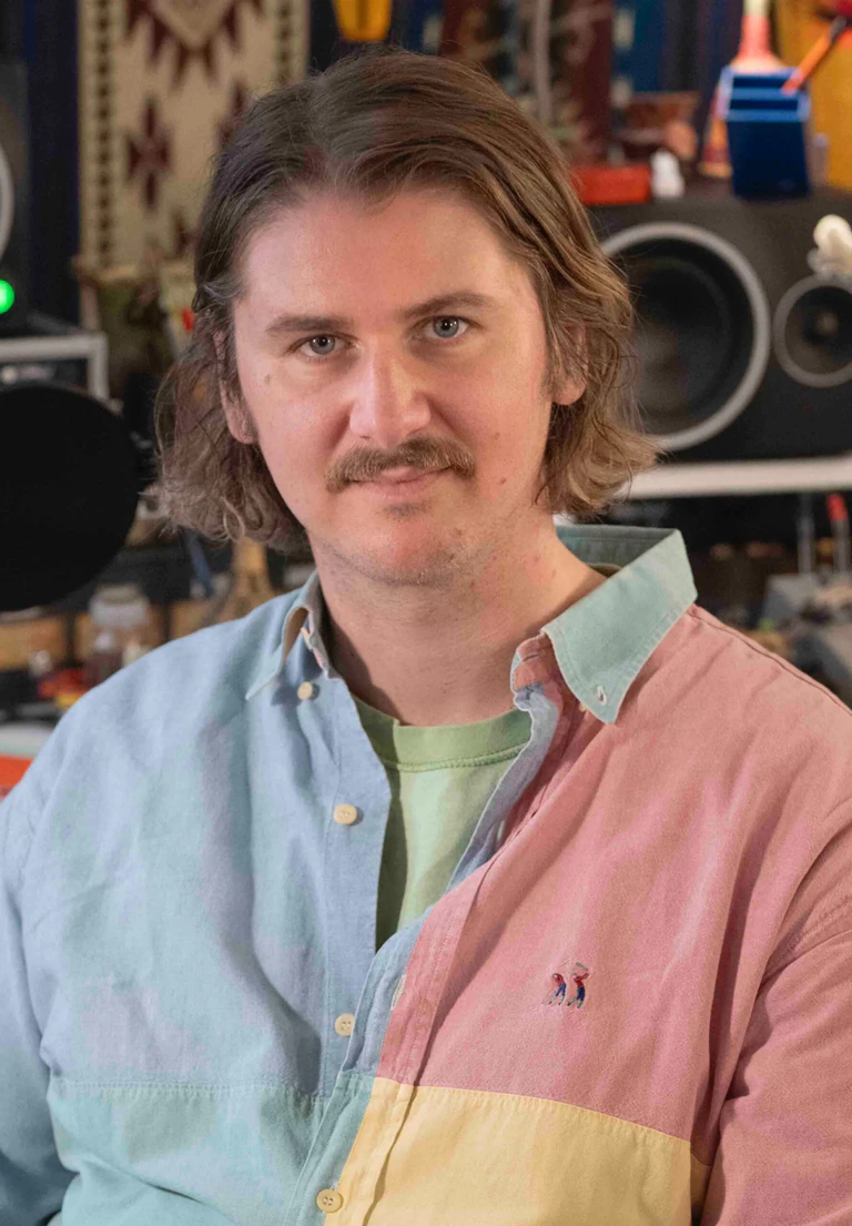 Portrait de Thibaud Vanhooland, alias Voyou, cheveux mi-longs et fine moustache, en chemise bicolore rose et bleue, dans son studio d'enregistrement