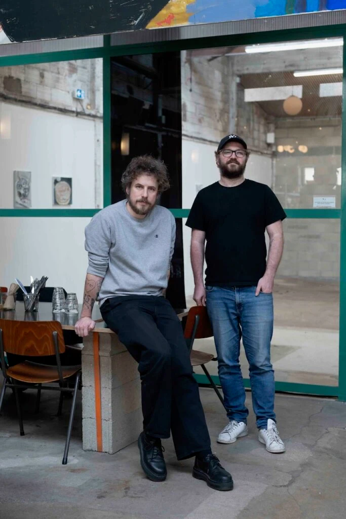 Deux hommes barbus posant devant les baies vitrées à cadre vert du restaurant, l'un assis sur un muret en parpaings, l'autre debout, la main dans la poche