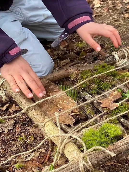 Un enfant accroupi en forêt manipule des branches et de la mousse autour d’un petit abri construit avec des éléments naturels.