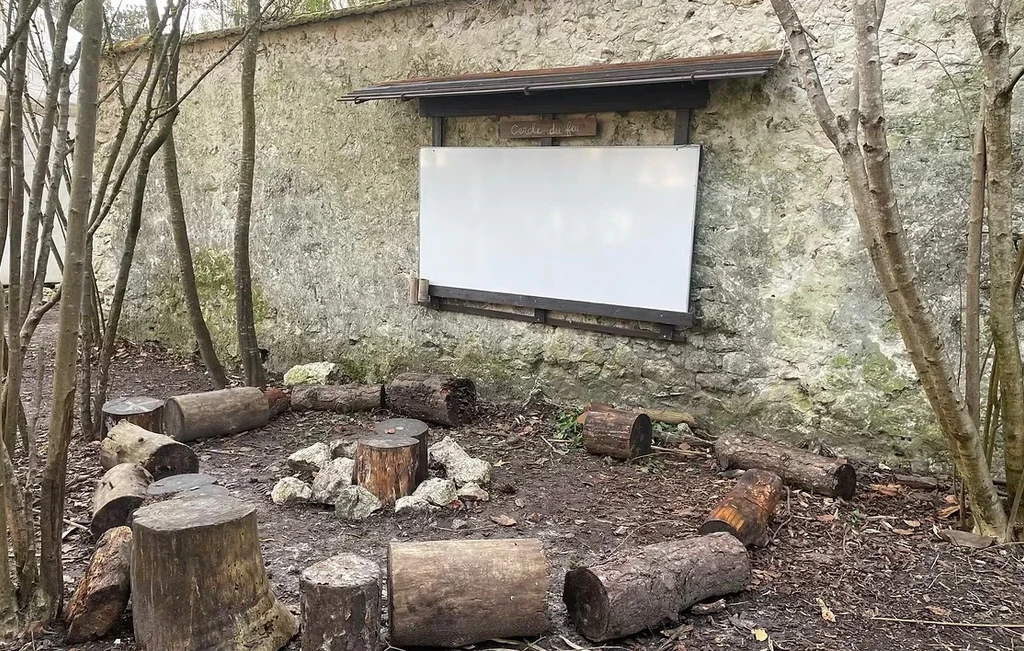 Espace pédagogique en pleine nature avec des rondins de bois disposés en cercle autour d’un foyer, face à un tableau installé contre un ancien mur en pierre.