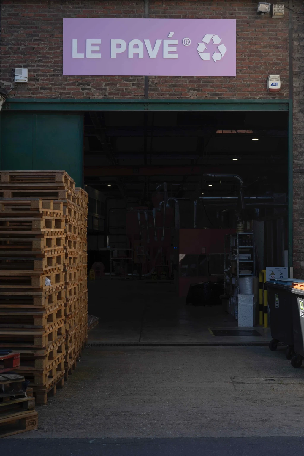 Façade en brique d'un entrepôt avec l'enseigne violette « Le Pavé » et son logo d'économie circulaire, palettes en bois à l'entrée.