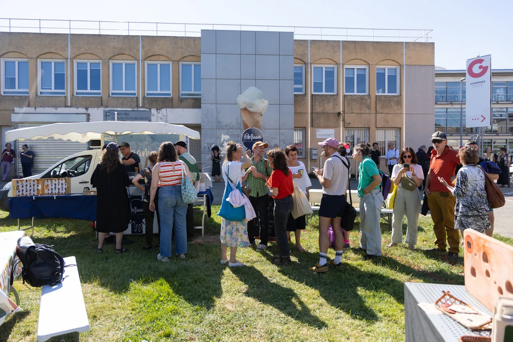 Des visiteurs font la queue devant plusieurs stands installés sur une pelouse. Au centre, un vendeur portant un chapeau sert des glaces depuis un stand mobile.