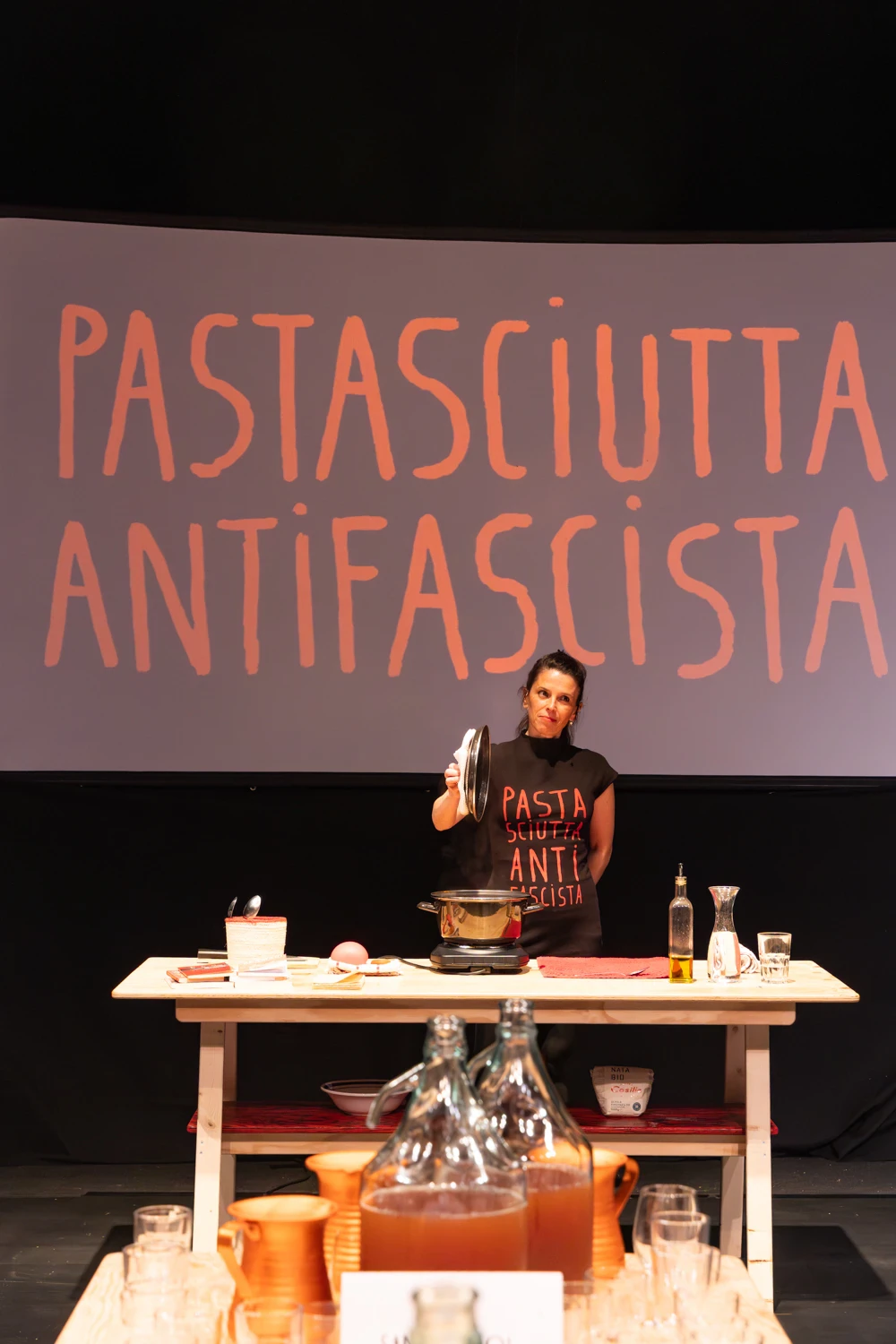 Femme vêtue de noir sur scène préparant une casserole de pâtes avec en arrière plan un écran reprenant le jeux de mots "Pastascuitta Antifascita"