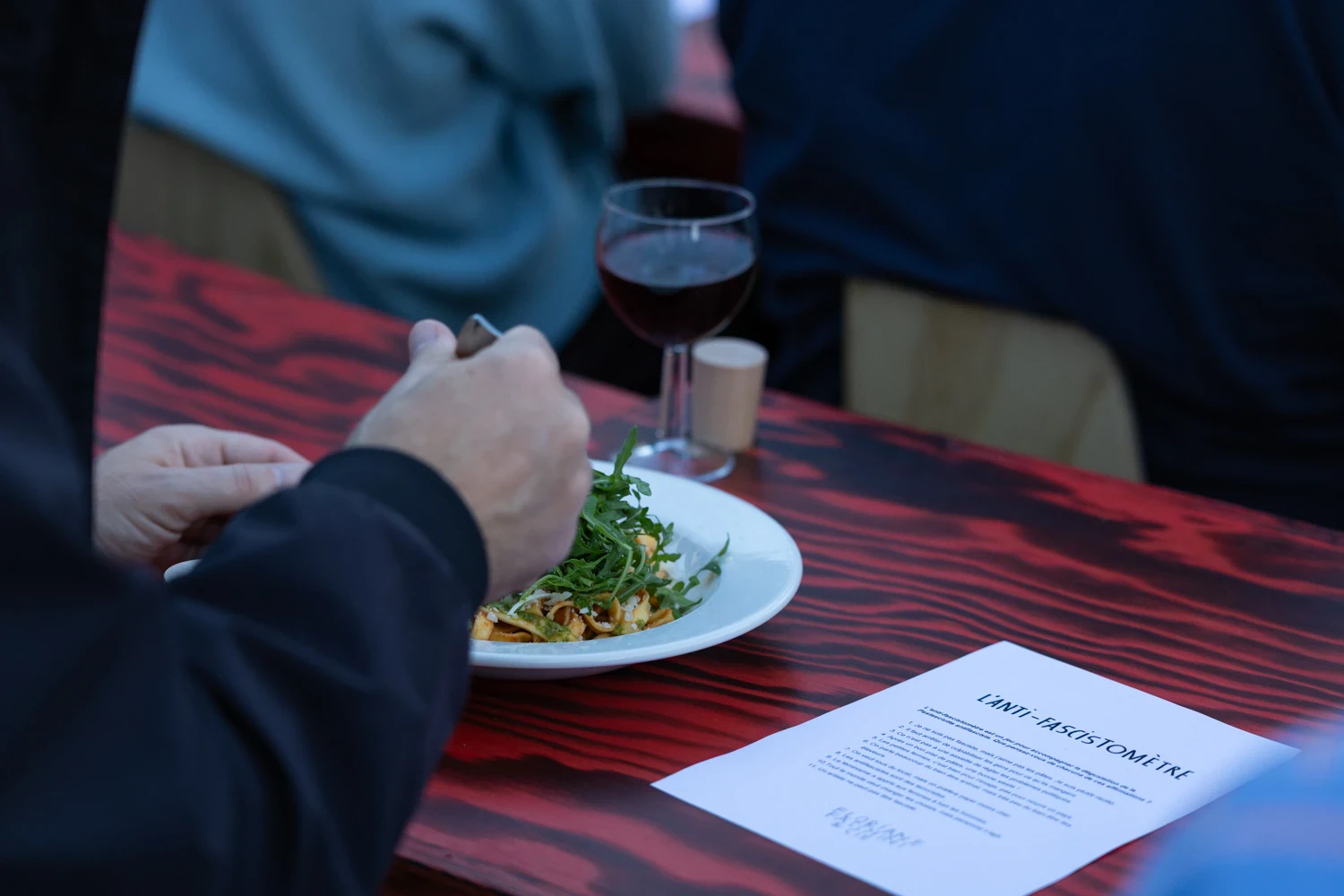 Une personne mange un plat de pâtes autour d’une table en bois rouge et noir. À côté de l’assiette, une feuille de papier portant le titre « L’ANTIFASCISTOMÈTRE » est posée sur la table, avec un verre de vin.