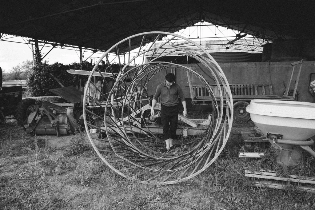 Photo en noir et blanc d'un homme marchant au centre d'une grande structure circulaire en bois, dans une cour de ferme entourée de matériel agricole
