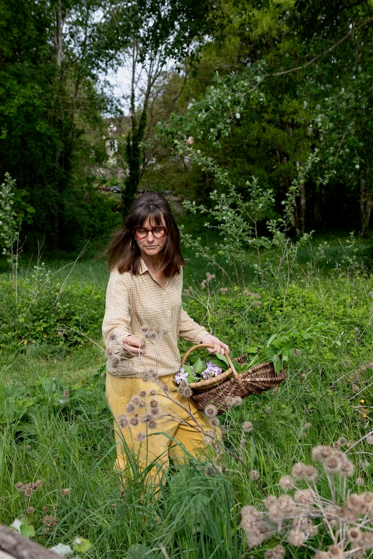 Femme aux cheveux bruns et lunettes, vêtue de jaune, traversant une prairie avec un panier rempli de plantes et de fleurs