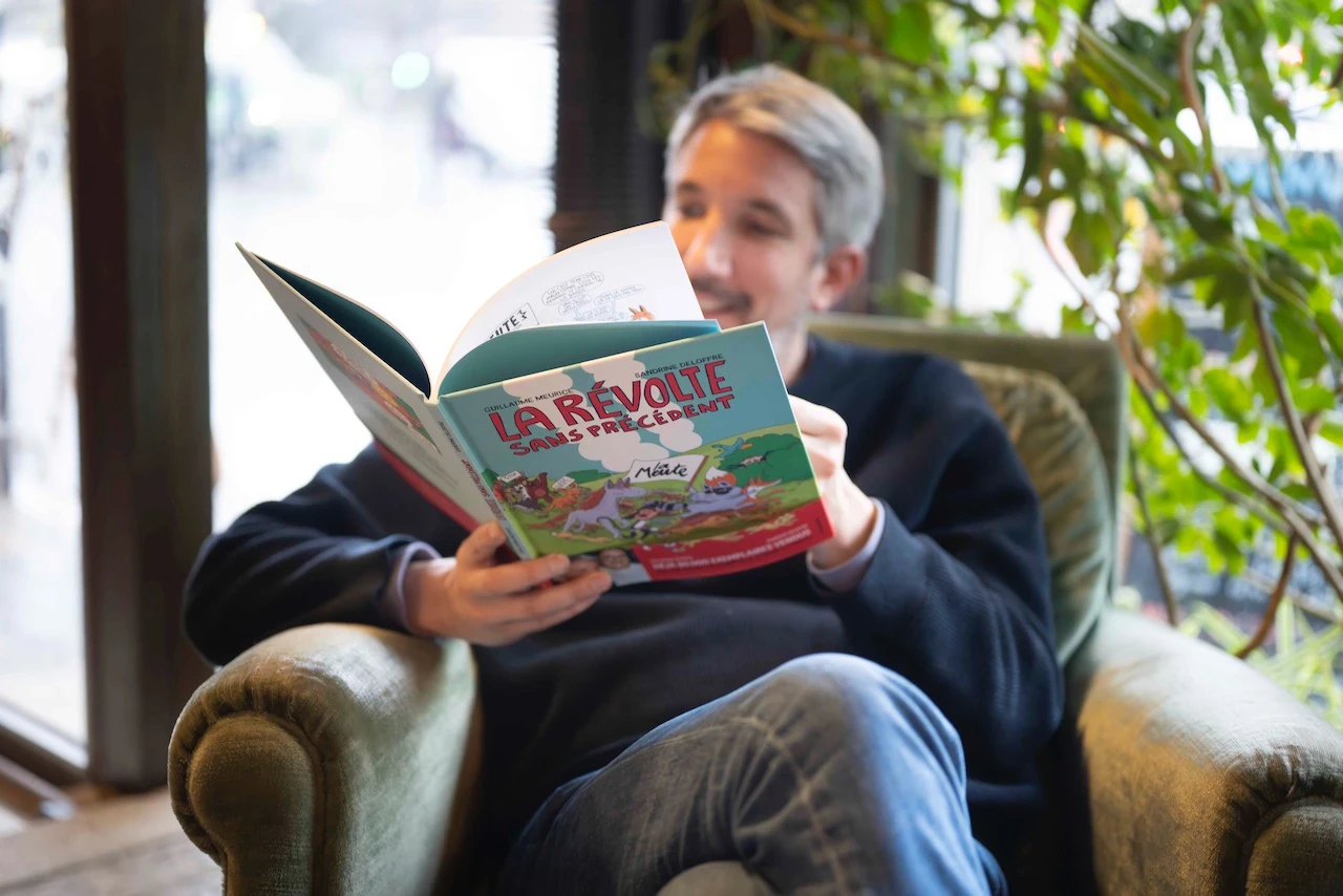 Portrait de guillaume Meurice, assis sur un fauteuil, lisant la bande dessinée La révolte sans précédent