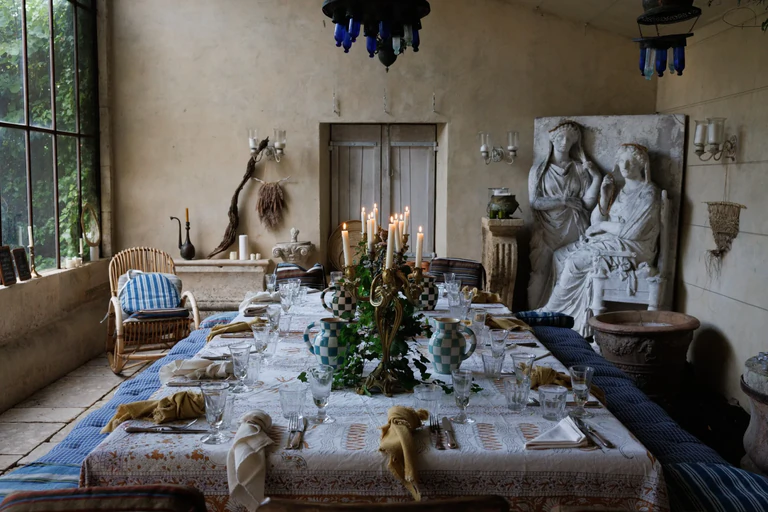 Grande table de réception dressée, avec nappe blanche et chandeliers allumés, dans une salle richement décorée (meubles en osiers, bas relief style romain et lustre imposant 