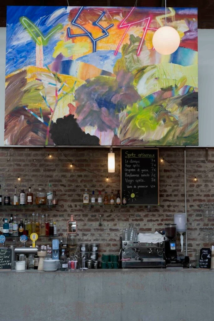 Comptoir de bar en béton devant un mur de briques, avec bouteilles, machine à café et ardoise de spritz, surmonté d'une grande peinture abstraite aux couleurs vives