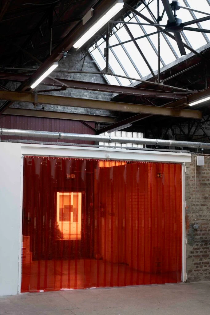 Grande salle industrielle à charpente métallique et verrière, avec une ouverture fermée par un rideau de lanières de plastique orange translucide