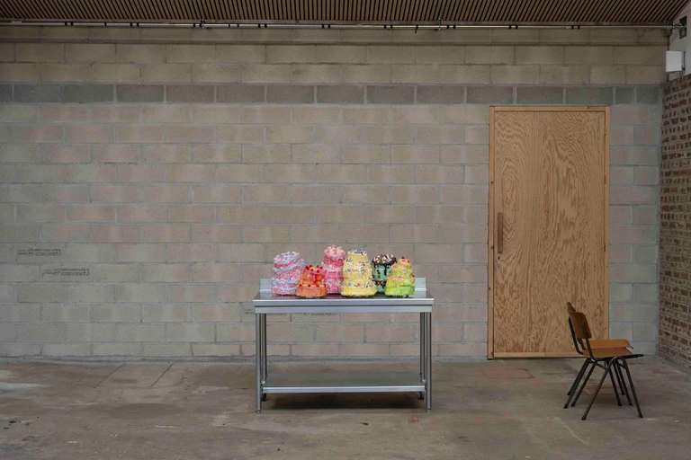 Sur une table en inox devant un mur en parpaings, une rangée de petits gâteaux sculptés roses, jaunes, verts et orange, à côté d'une porte en contreplaqué et d'une chaise d'écolier