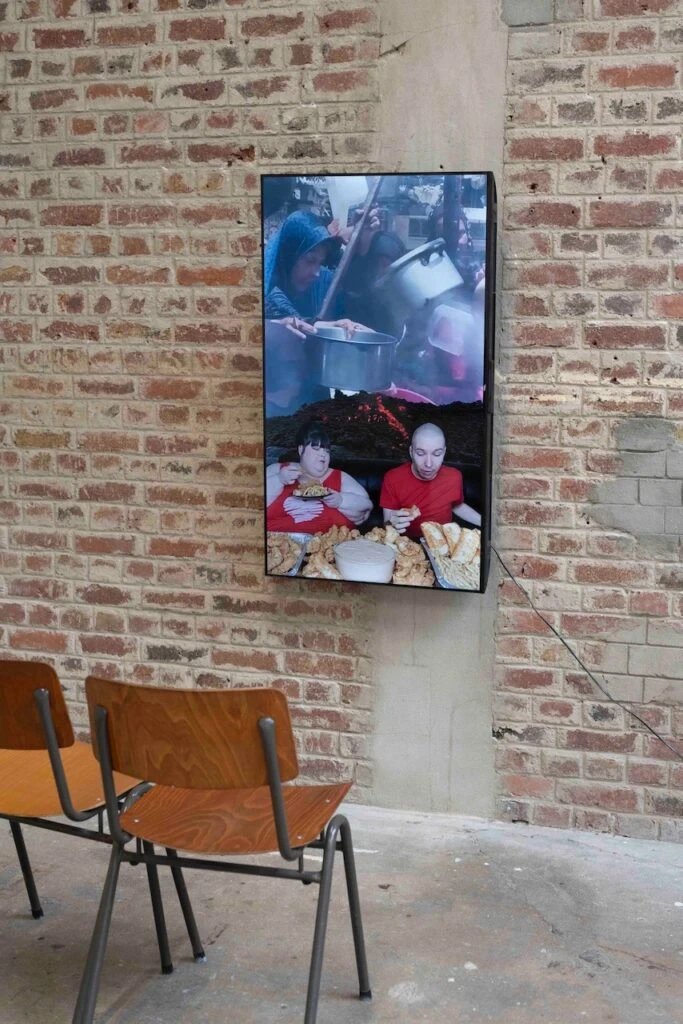 Écran vertical accroché à un mur de briques, montrant deux personnes attablées devant des plats et, au-dessus, des mains tendant des casseroles, avec deux chaises en bois devant