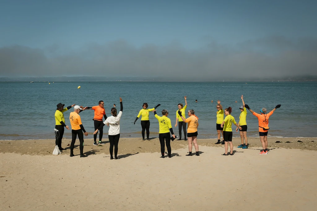 Groupe d'une dizaine de participants, debout sur la plage, font tourner leurs bras en guise d'échauffement