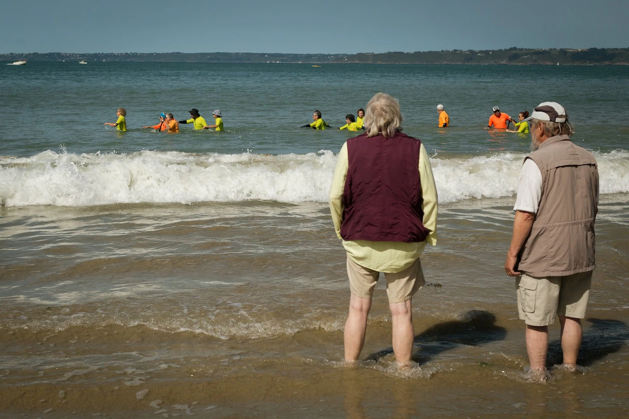 Deux hommes les pieds dans l'eau, la cinquantaine, observent depuis la plage les pratiquants de longe côte