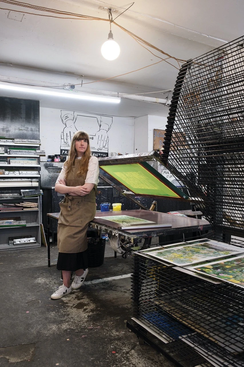 Portrait de Lucille Clerc, en tablier, debout à côté d'une presse à sérigraphie dans son atelier encombré de tirages