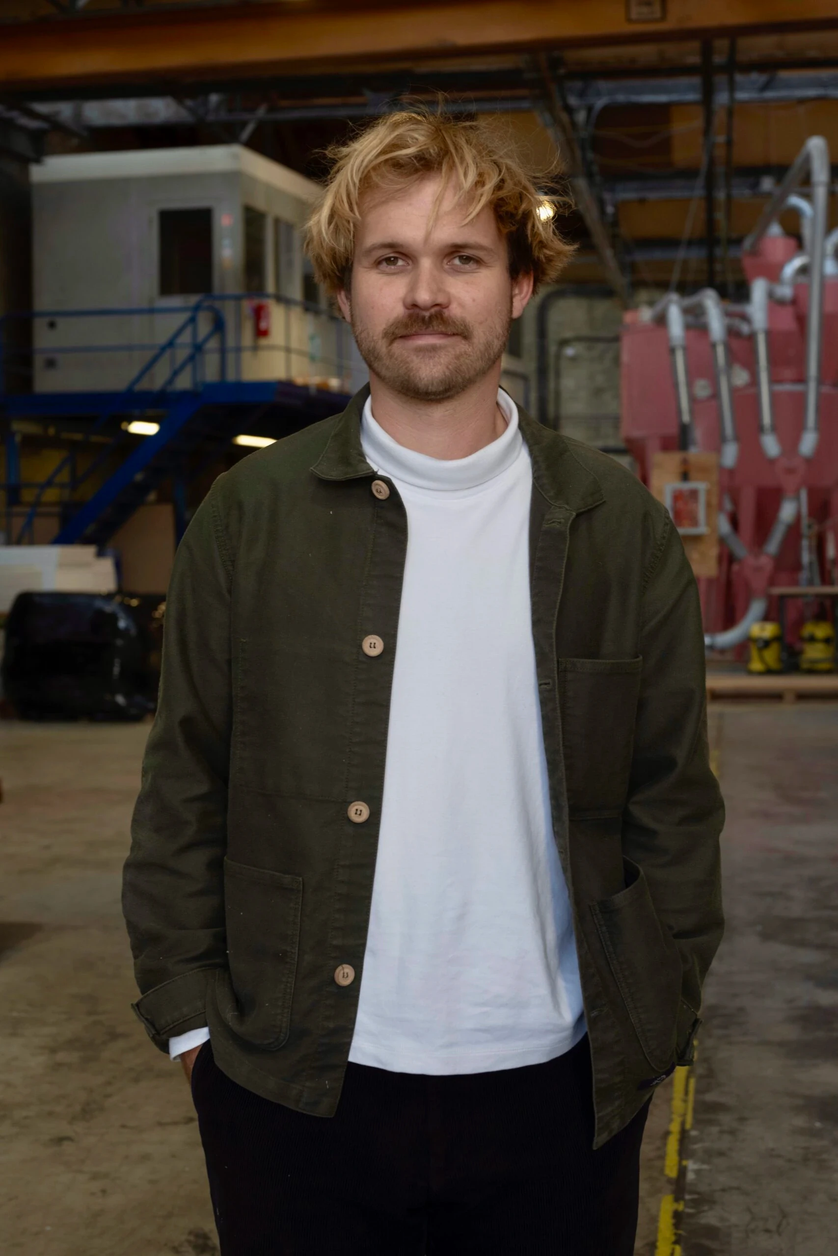 Portrait de Marius Hamelot, veste kaki et pull blanc, debout dans l'usine industrielle Le Pavé.