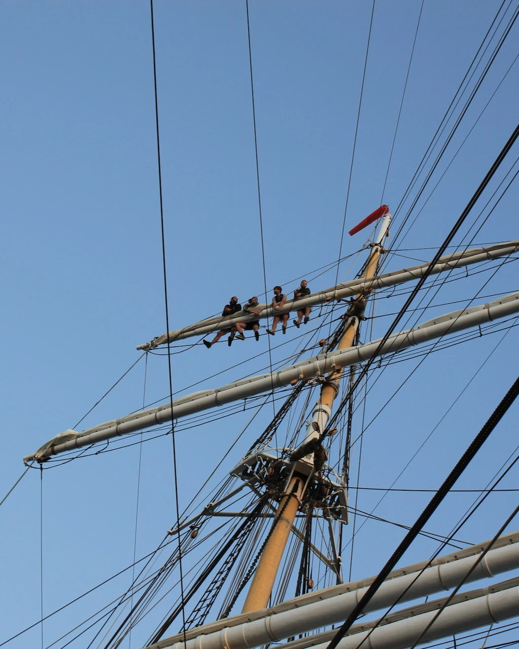 Quatre marins assis en équilibre sur une vergue, en haut du mât d'un grand voilier, ciel bleu dégagé