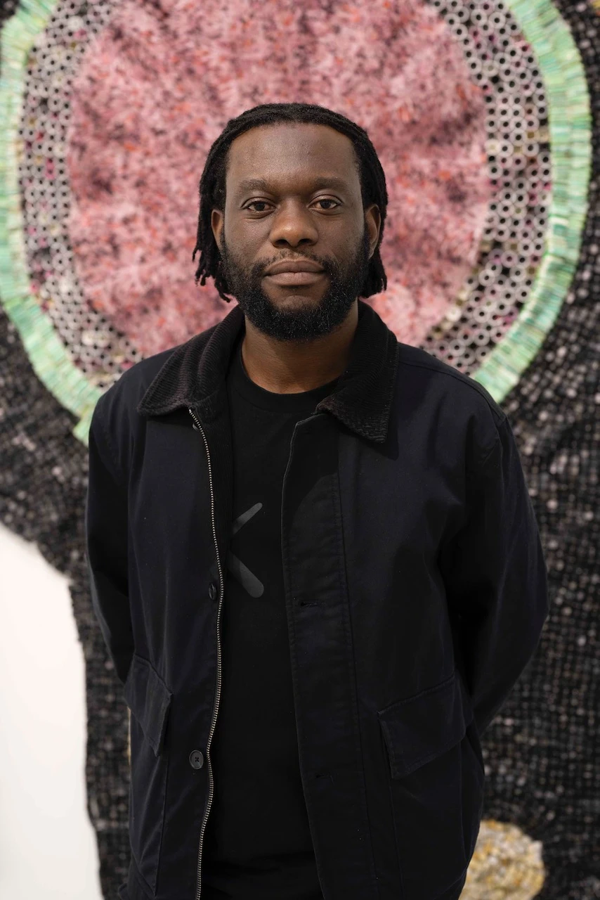 Portrait de Moffat Takadiwa, vêtu de noir, posant devant une tapisserie rose et verte