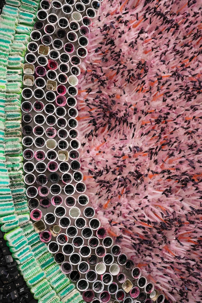 Gros plan sur une œuvre textile faite d'embouts de tubes assemblés en cercles, à côté de brosses à dents roses et orange