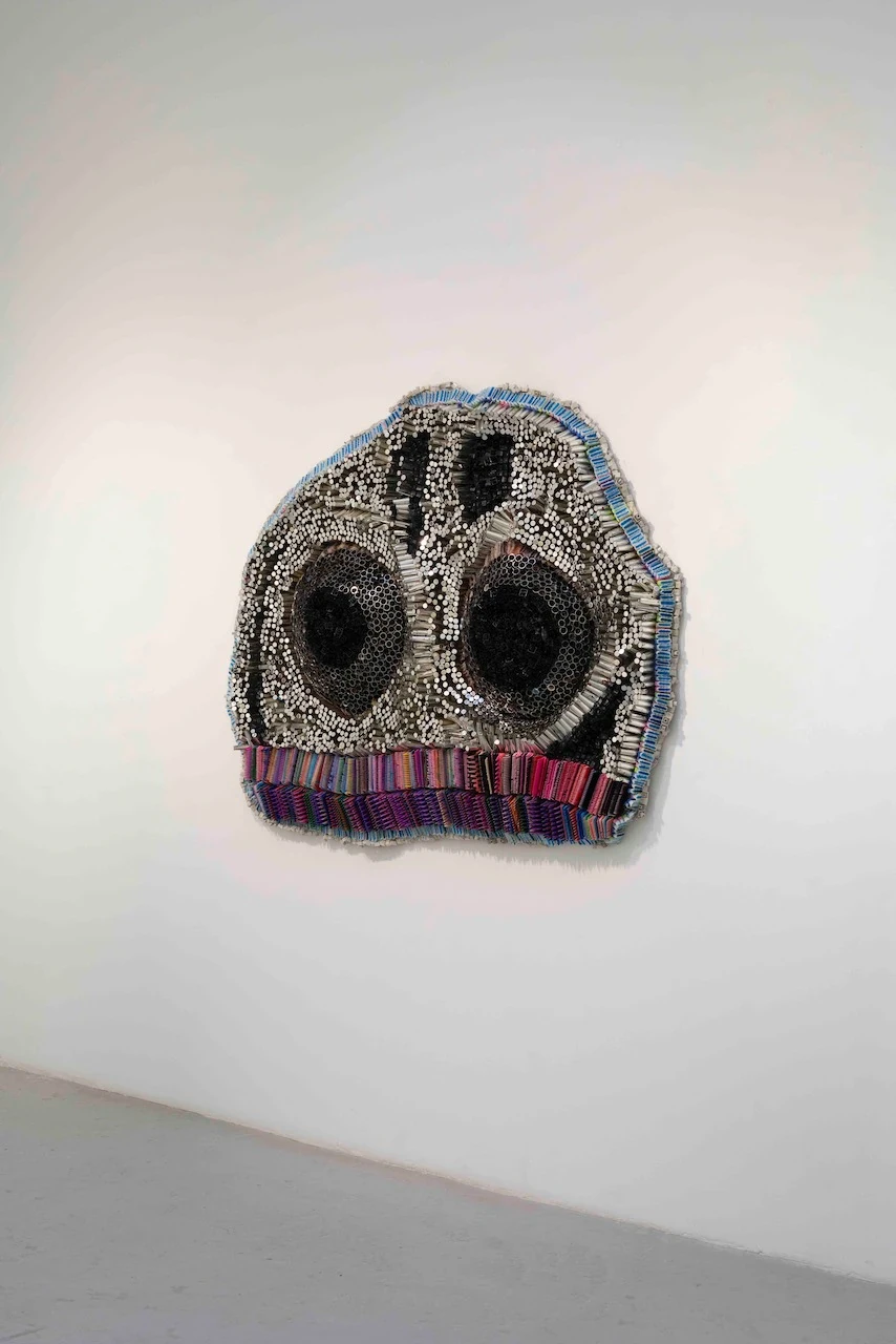 Sculpture murale évoquant un visage aux grands yeux noirs, faite de tubes métalliques et de bandes textiles colorées