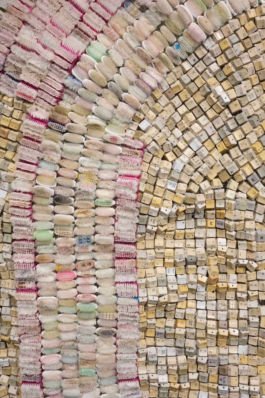 Gros plan sur une tapisserie textile mêlant brosses à dents usagées roses et vertes et touches de clavier d'ordinateur beiges