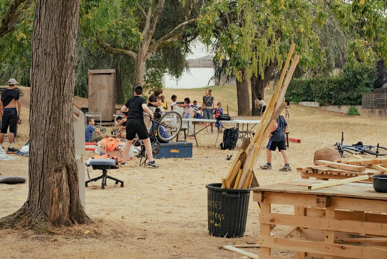 Terrain en herbes avec au premier plan un établi fait en cagette de bois et une pile de planches. En arrière plan un groupe d'enfants et d'adultes jouent ou discutent