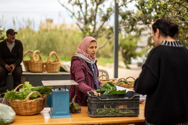 Jeune femme voilée en rose derrière une table en bois garnie de paniers de légumes verts, d'une balance bleue et d'une caisse de feuilles, face à une cliente de dos