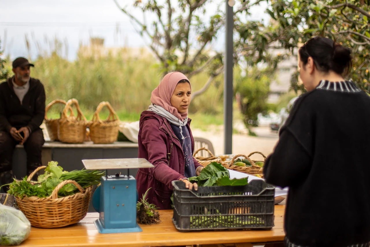 Jeune femme voilée en rose derrière une table en bois garnie de paniers de légumes verts, d'une balance bleue et d'une caisse de feuilles, face à une cliente de dos