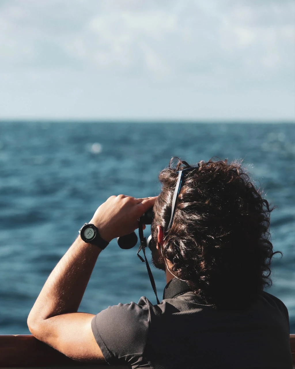 Homme de dos observant l'océan aux jumelles, cheveux bouclés au vent, en mer.