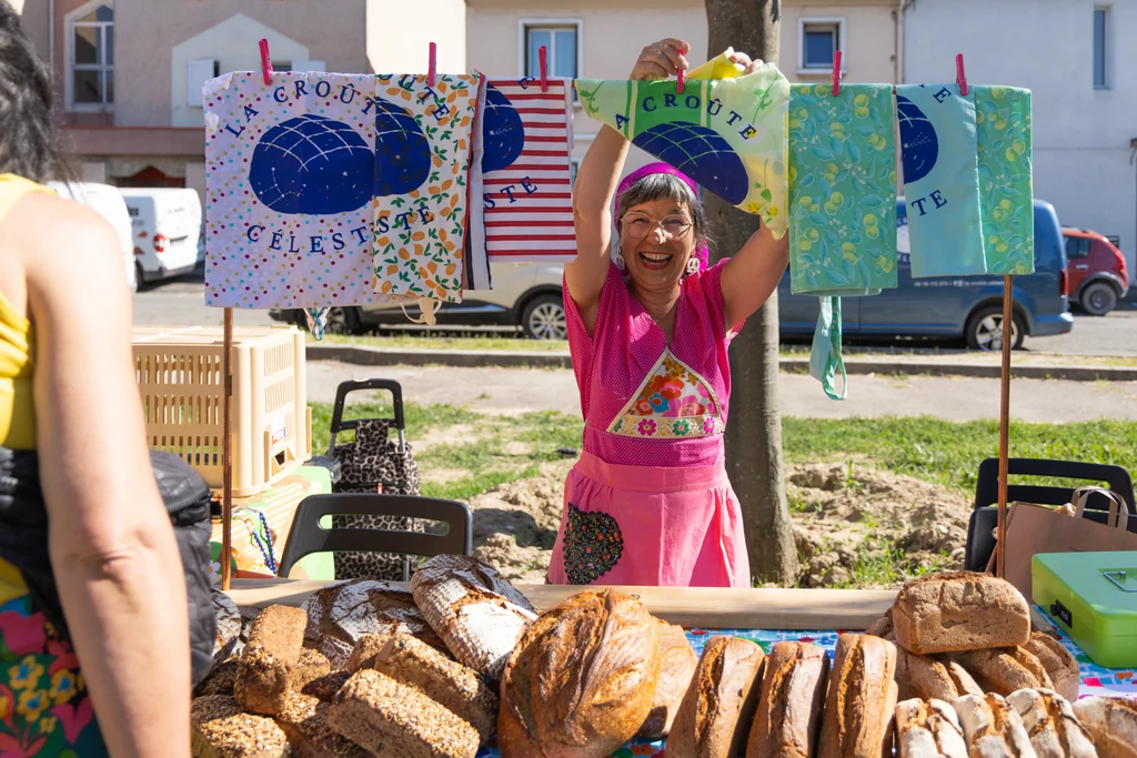 Une boulangère souriante, vêtue d’un tablier rose, présente des pains et des produits de boulangerie sur un stand en plein air. Des tissus colorés sont suspendus au-dessus d’elle.