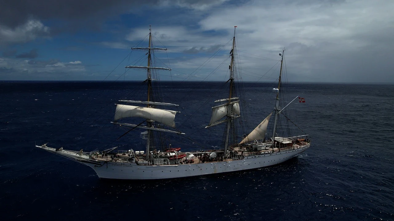 Grand voilier trois-mâts naviguant en pleine mer, voiles à moitié déployées, sous un ciel nuageux